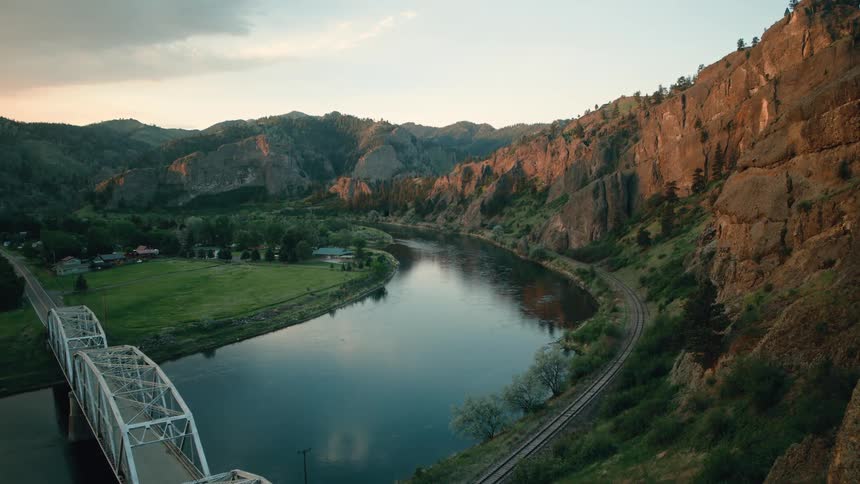 An aerial view slowly moves towards a steel bridge crossing a wide river nestled in a rocky mountain canyon at sunset