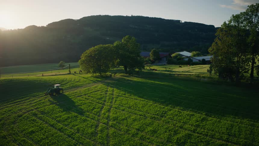Aerial footage shows a tractor working in a lush green field on a rural farm as the golden sun sets behind rolling hills