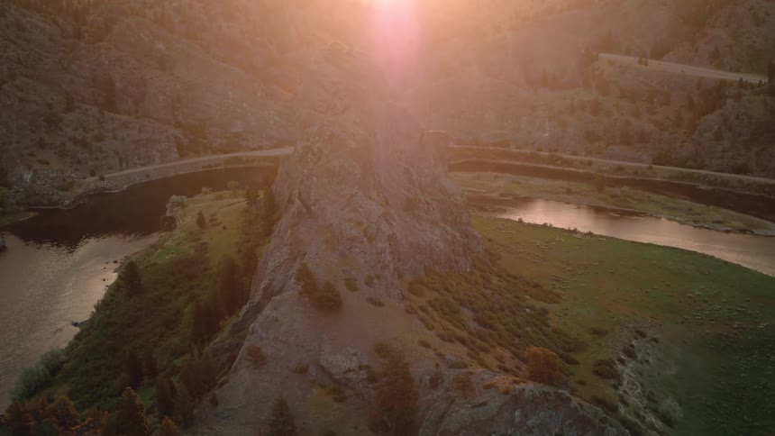 An aerial view rises over a majestic rocky mountain peak as the golden sun sets, revealing a winding river below