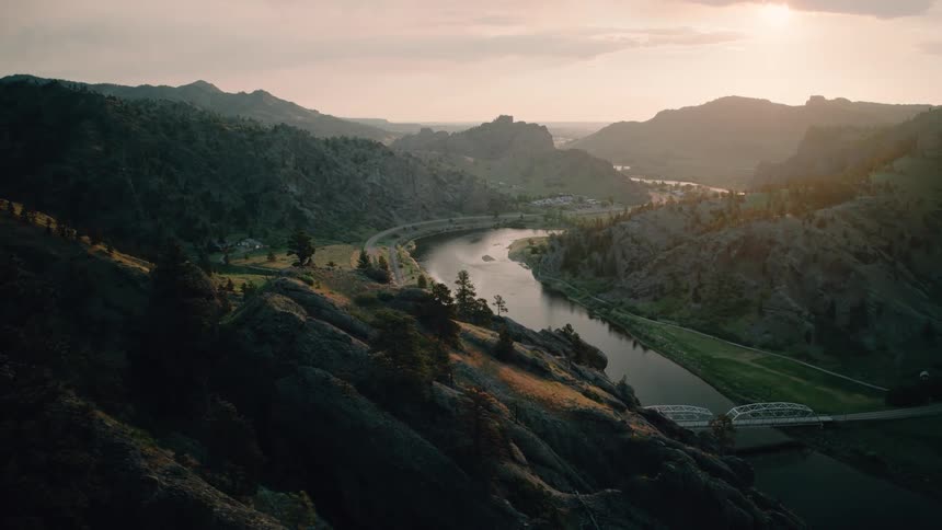 A slow aerial pullback shot over a scenic mountain valley showcases a winding river and a highway glowing in the warm sunset light
