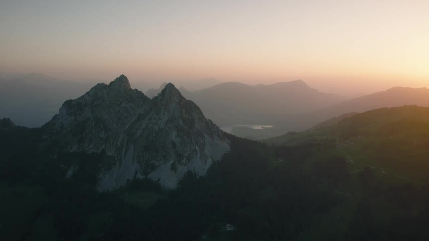 A serene aerial view shows majestic mountain peaks and a lush valley with a distant lake during a hazy golden hour sunrise