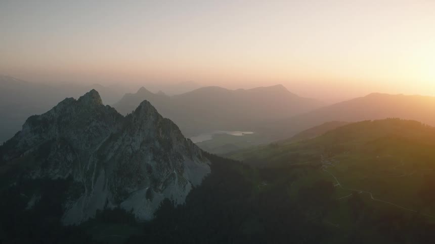 A static aerial view captures the majesty of a mountain range with a lake in the valley during a hazy, golden sunrise