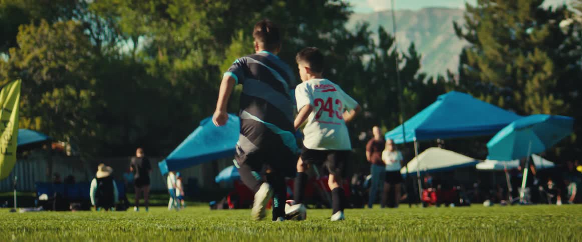 A dynamic ground-level shot follows a group of young boys as they dribble and chase the ball during a competitive soccer game on a sunny day