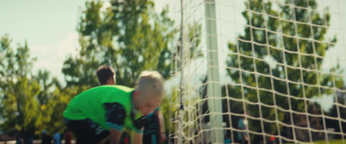 view from behind the net as young boys play a soccer match and a player retrieves the ball after it goes out of bounds