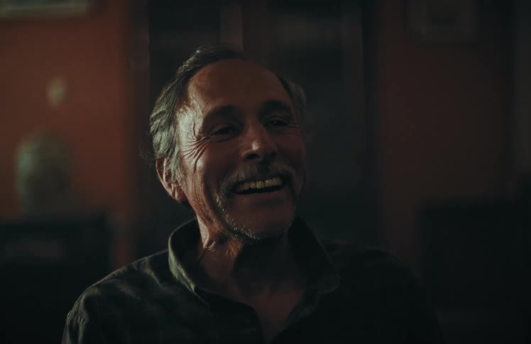 A joyful elderly man with a mustache laughs while sitting in a dimly lit room