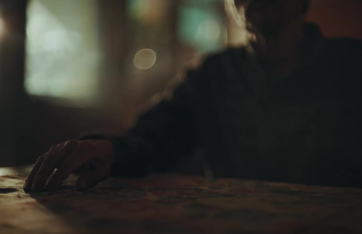 A man's hand rests on a table in a dimly lit room, creating a moody and suspenseful atmosphere