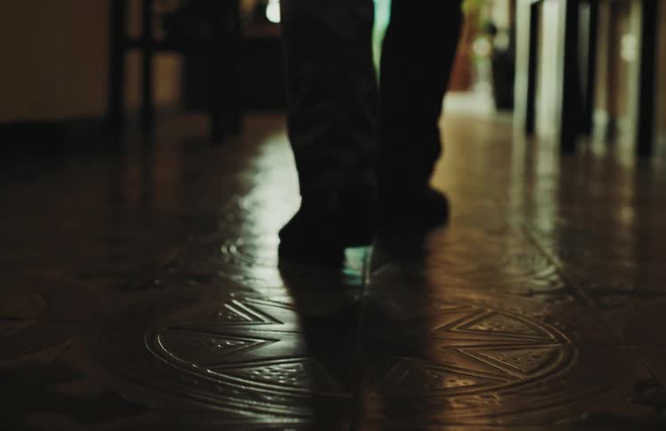 A mysterious silhouetted figure walks away in slow motion on a reflective, patterned tile floor