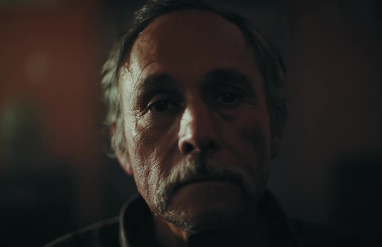 A dramatic close-up portrait of a serious senior man with a mustache turning his head to look into the camera in a dark, moody room