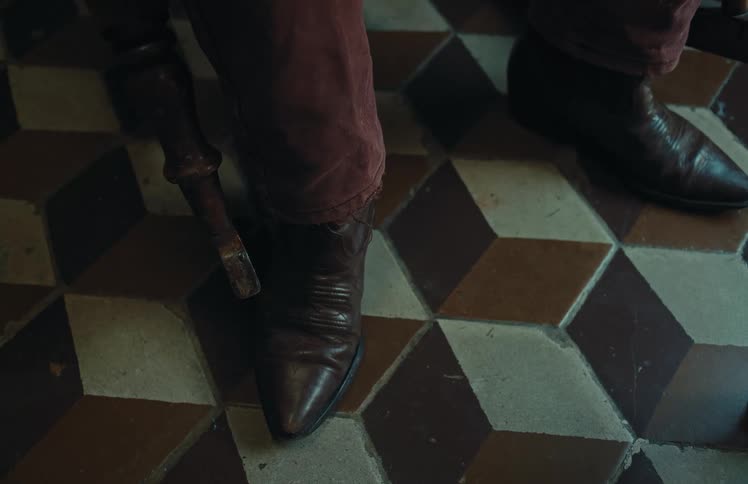 A static close-up shot shows a person's brown leather boots resting on a vintage geometric tiled floor