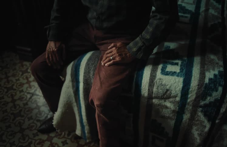 A pensive man with a wedding ring sits on a patterned blanket in a dimly lit bedroom, his hands resting on his legs