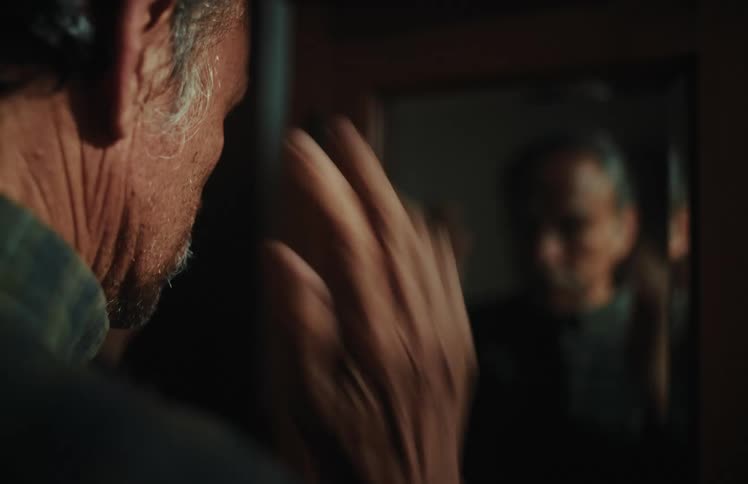 An over-the-shoulder view of a senior man thoughtfully getting ready in front of a mirror in a dimly lit room