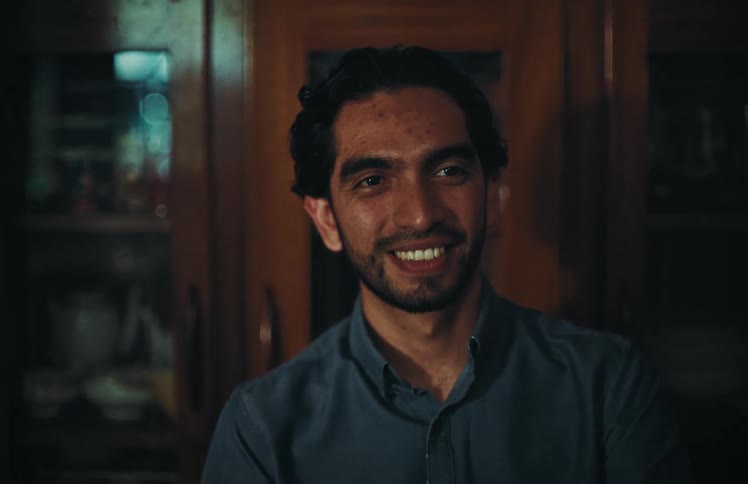 A handsome young man with a beard laughs heartily and then settles into a warm smile in a dimly lit room