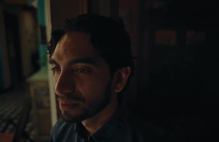 A moody close-up shot captures a pensive young man looking off-camera in a dimly lit room