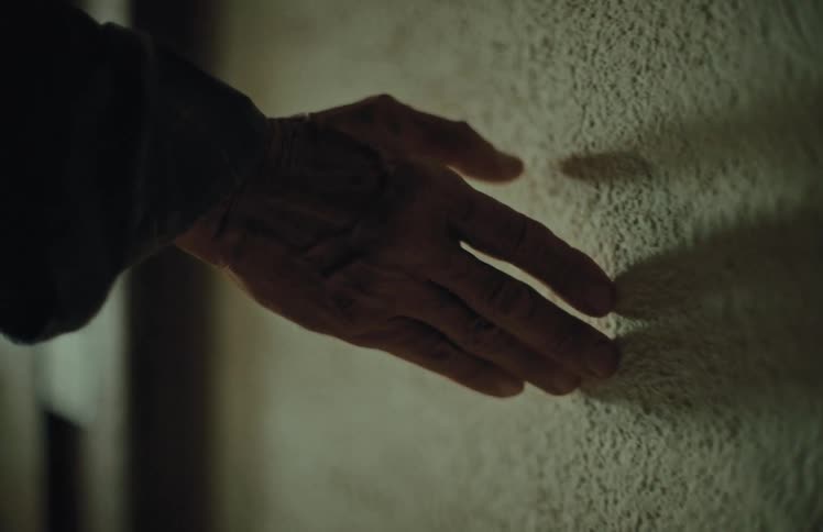 A close-up of a senior's hand gently sliding across a rough textured wall in a dimly lit space