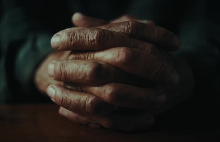 An anxious elderly person wrings their weathered hands in a dramatic, low-light close-up