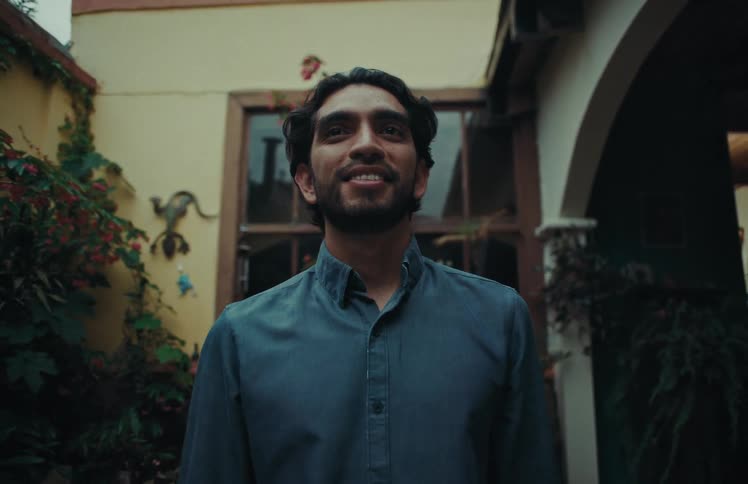 A handsome, smiling man walks towards the camera in a lush, green courtyard