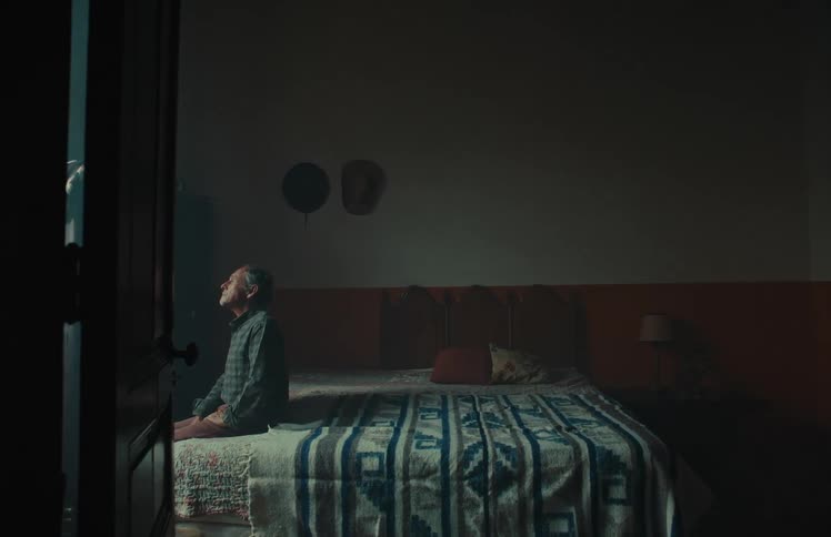 A pensive elderly man sits alone on the edge of his bed in a dimly lit, rustic bedroom, looking thoughtfully towards the light