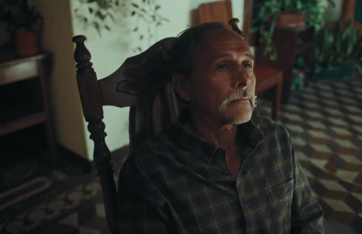 A contemplative older man with a mustache opens his eyes while sitting in a wooden rocking chair inside his home