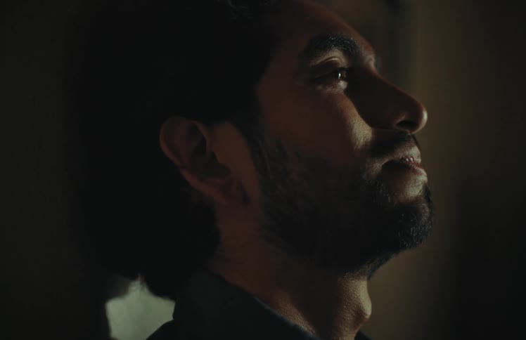 A pensive man with a beard opens his eyes in a dramatically lit room, suggesting a moment of realization or hope