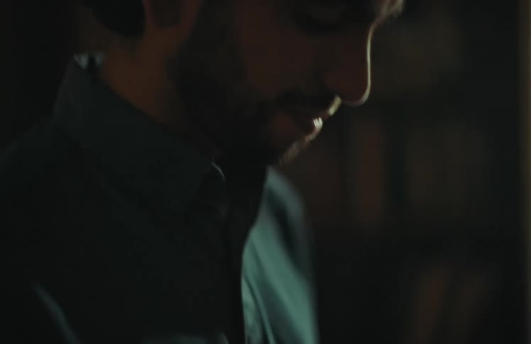 A somber man with a beard looks down at an old, black and white photograph in a dimly lit room