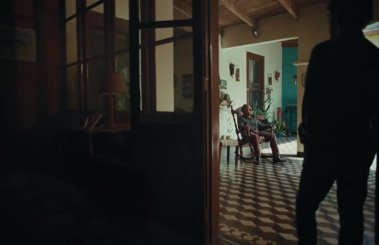 A silhouetted man stands in a doorway, observing an older man relaxing in a rocking chair inside a sunlit, rustic house