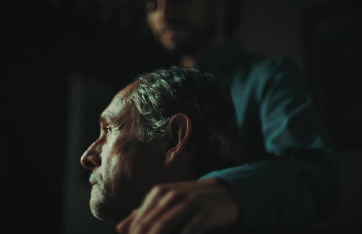 An adult son offers comfort and support to his elderly father, placing a reassuring hand on his shoulder in a dimly lit room