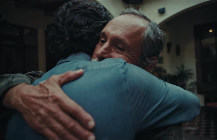 An elderly father and his adult son share a heartfelt, comforting hug in a dimly lit courtyard