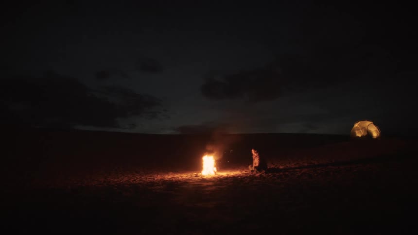 A lone figure sits by a crackling campfire in the vast desert at night, with an illuminated tent in the background