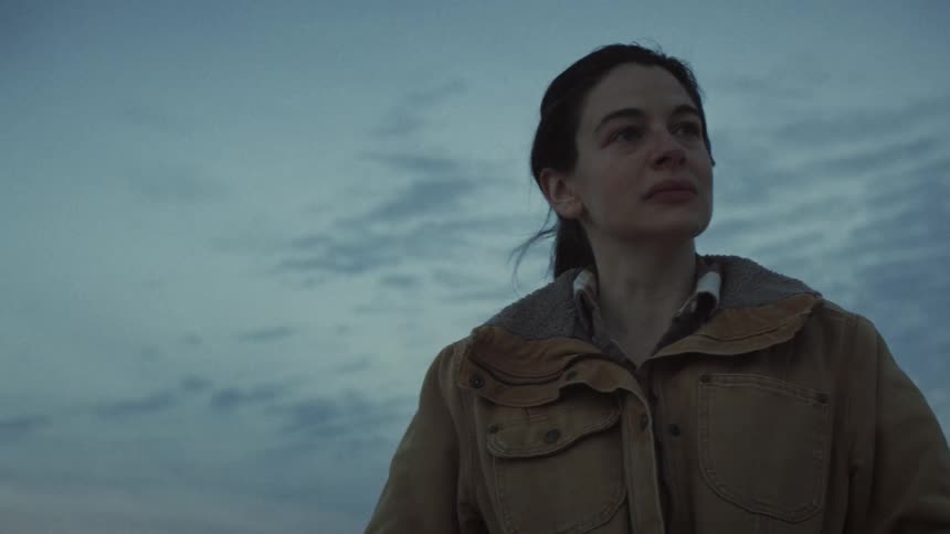 A low-angle shot of a woman with a tearful, bittersweet expression standing against a vast, gloomy sky