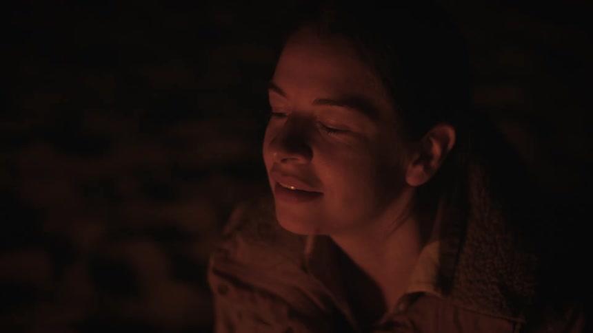 A serene woman smiles softly as her face is illuminated by the warm, flickering light of a campfire at night