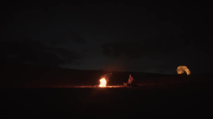 A lone camper tends a bright campfire in the middle of a vast, dark landscape at night, with a glowing tent in the distance