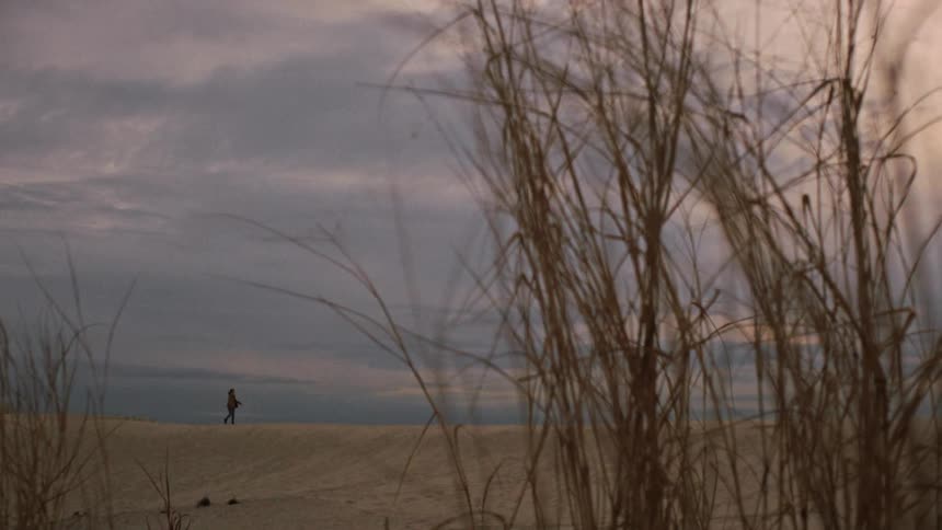 A solitary figure walks and spins freely along the horizon of a vast sand dune under a cloudy twilight sky, viewed from behind tall beach grass