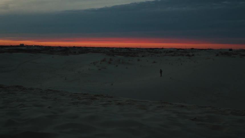 A lone figure stands amidst vast, shadowy sand dunes against the backdrop of a fiery red sunset on the horizon