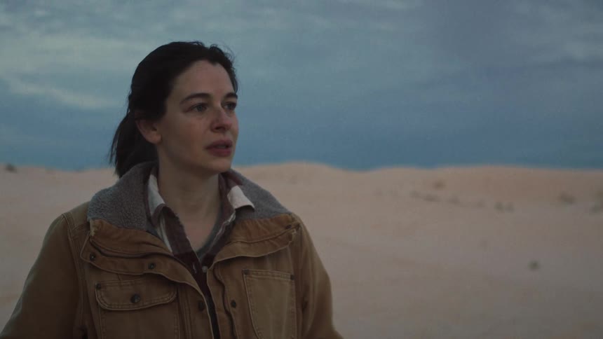 A woman with a pensive expression stands alone in a vast, windy desert at dusk