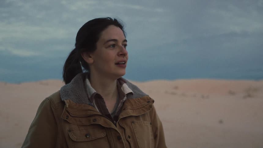 A thoughtful woman stands alone in a vast desert landscape at dusk, a subtle smile of hope giving way to a more somber expression