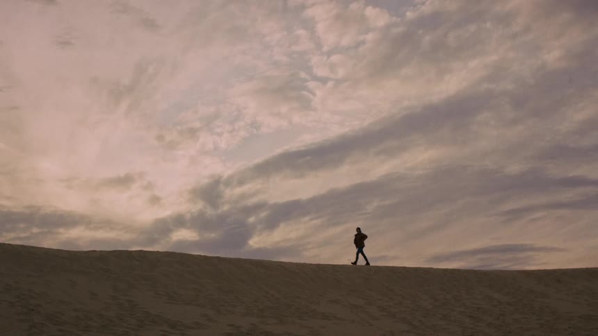 A lone silhouetted figure walks along the ridge of a large sand dune under a vast, dramatic sky
