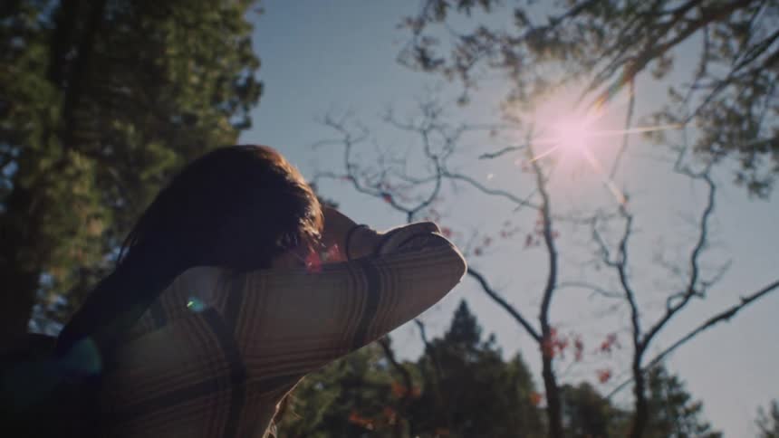 A woman hiker shields her eyes as she looks up at the bright sun shining through the tall forest trees in slow motion