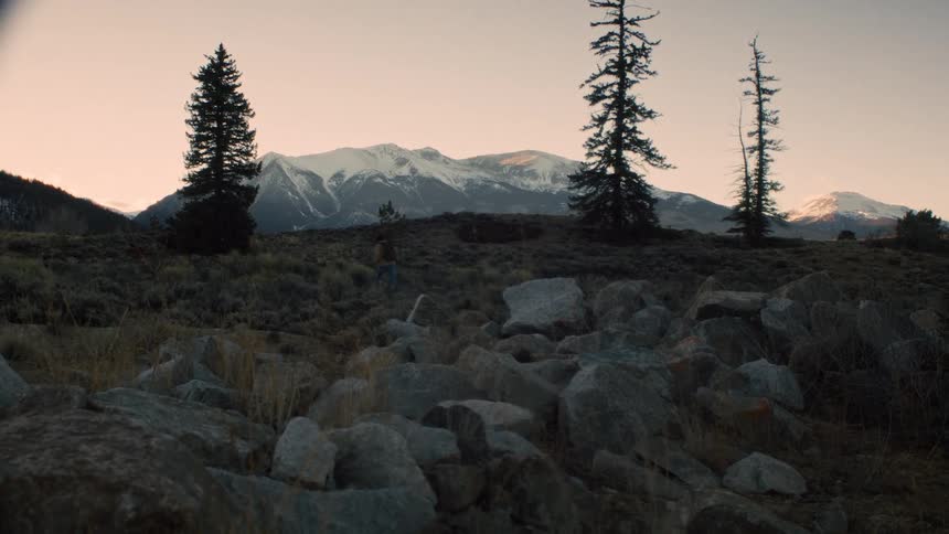 A lone hiker walks up a rocky, grassy hillside towards snow-capped mountains at dusk