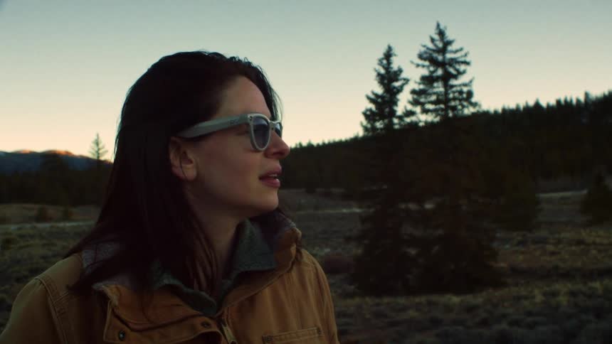 A thoughtful woman wearing sunglasses and a warm jacket gazes at the mountain scenery during a golden hour sunset in slow motion