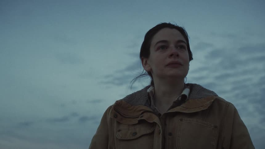 Close-up of a thoughtful and emotional woman in a work jacket looking up towards the overcast sky at twilight