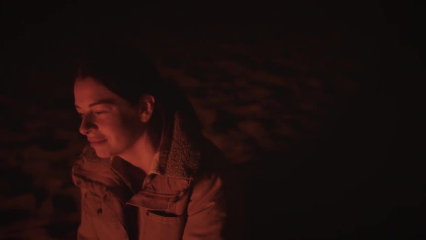 A serene woman smiles as she sits by a campfire at night, her face glowing in the warm light