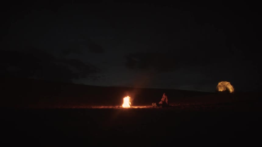 Wide shot of a person sitting by a campfire at night in the wilderness with an illuminated tent in the background