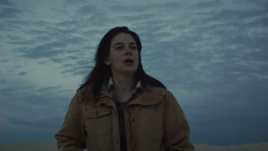 A pensive woman walks toward the camera in slow motion through a vast, desolate sand dune landscape at dusk