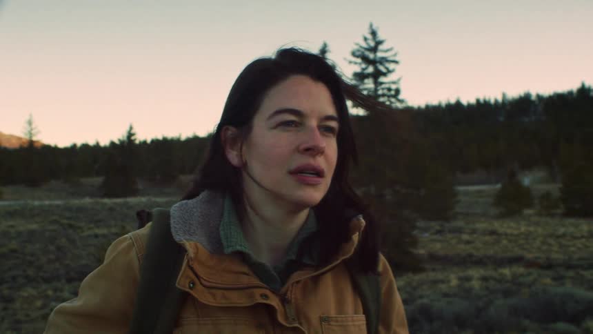 A pensive woman hiker pauses in a windy landscape at sunset before continuing on her journey in slow motion