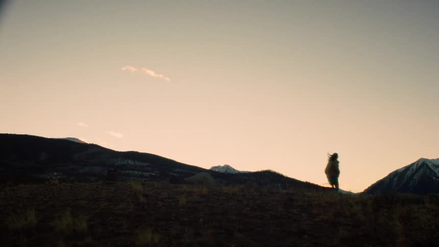 A silhouetted person joyfully walks and skips along a mountain ridge at sunset, eventually disappearing out of frame