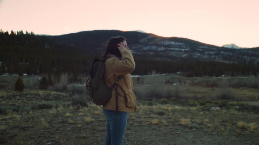 A female hiker with a backpack walks through a mountain pass at sunset, stopping to take in the beautiful scenery in slow motion