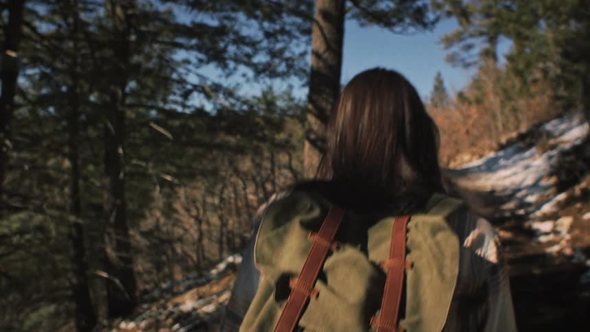 A woman with a backpack hikes a snowy mountain trail through a sunlit forest in slow motion