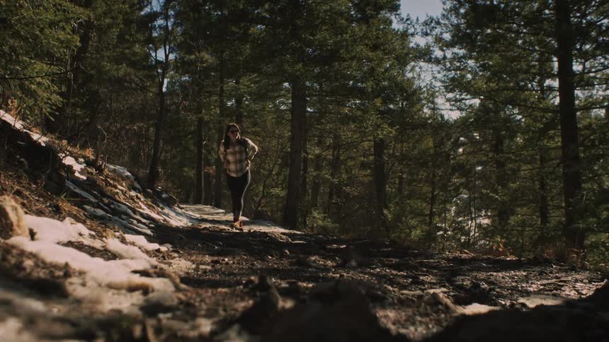 A woman pauses to enjoy the view before hiking down a muddy and snowy forest trail on a sunny day in slow motion