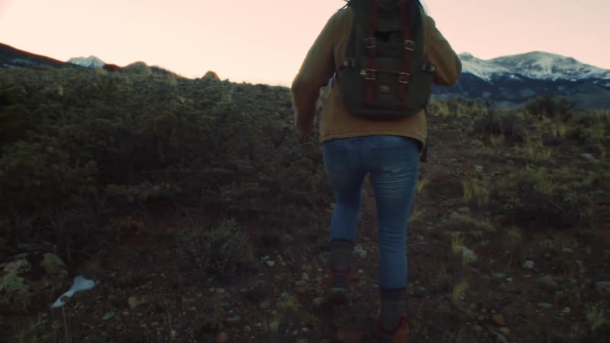 A woman with a backpack hikes up a mountain trail at sunset with snowy peaks in the distance