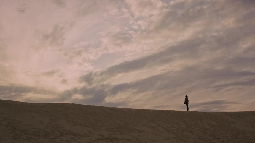 A lone figure stands silhouetted on a vast sand dune against a dramatic, cloudy sky